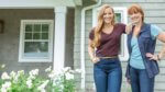 A mother/daughter pair in casual work clothes in front of a pretty suburban home