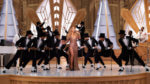 Mariah Carey on stage in a sparkling dress with a group of male dancers behind her