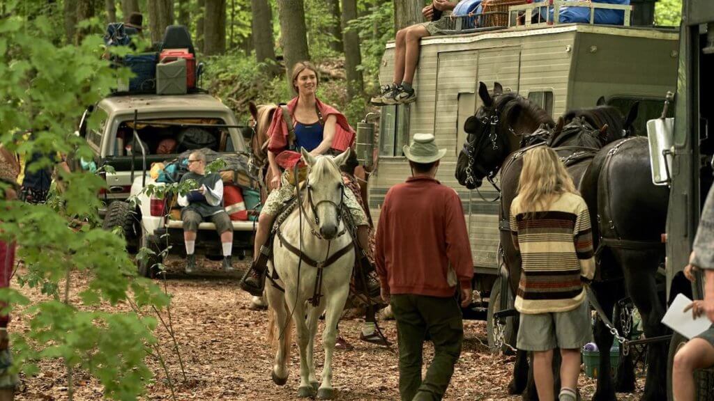 A woman rides a horse along a caravan of misc vehicles loaded with baggage