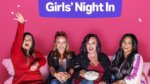 Four girls eat popcorn on a couch looking at camera with a pink background