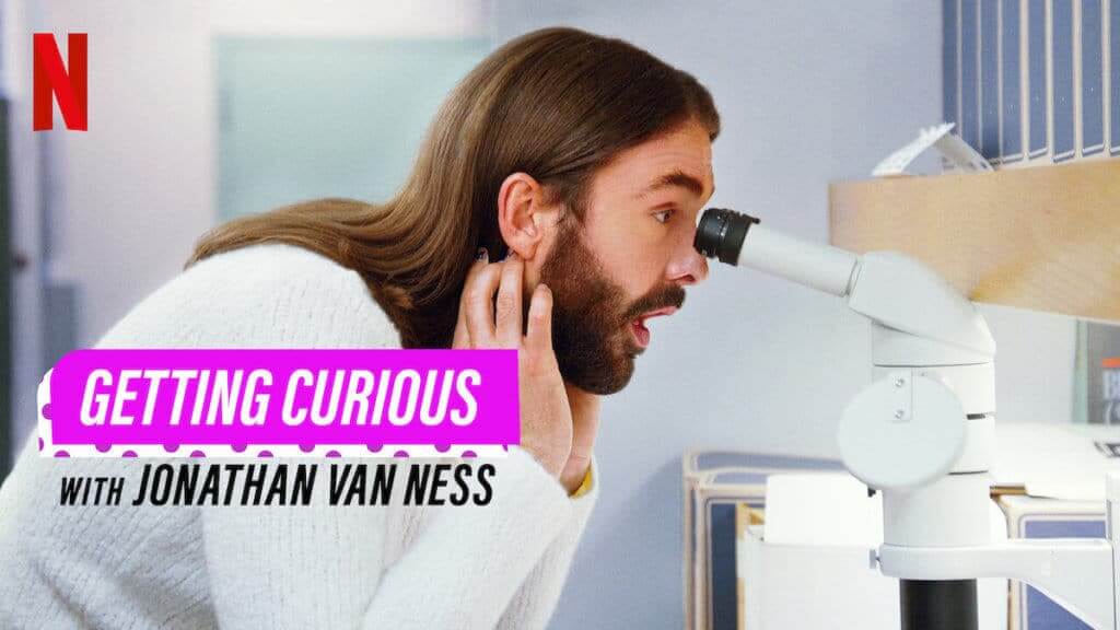 A person with long brown hair and a beard leans into a microscope with a surprised look on their face