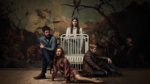 Four people posed eerily around an old fashioned crib in a creepy room