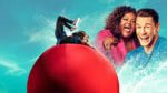 A person slamming into a big red ball with two hosts laughing behind them
