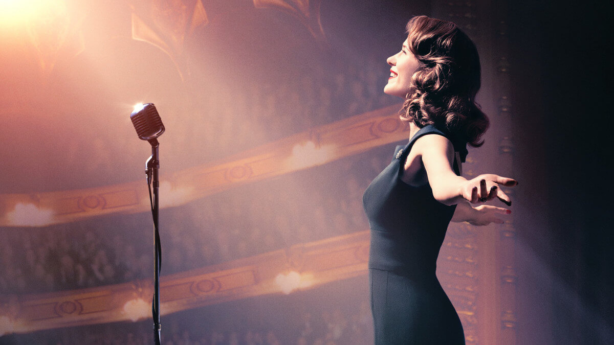 A woman in a black cocktail dress stands in profile with arms outstretched at a vintage microphone and in light of a spotlight