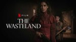 A woman stands in a shadowy cabin with a rifle and a boy behind her holding an ax