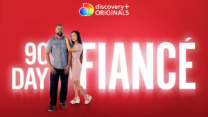 A couple stands in front of the show logo on a red background.