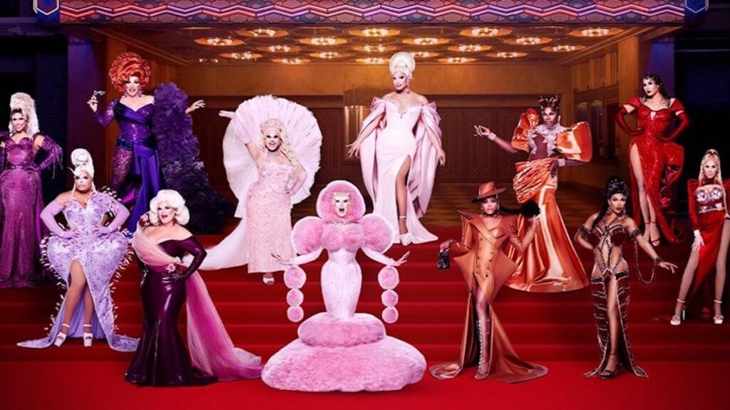 A group of fabulous drag queens on the steps of a theatre in elaborate pink, red, and purple costumes