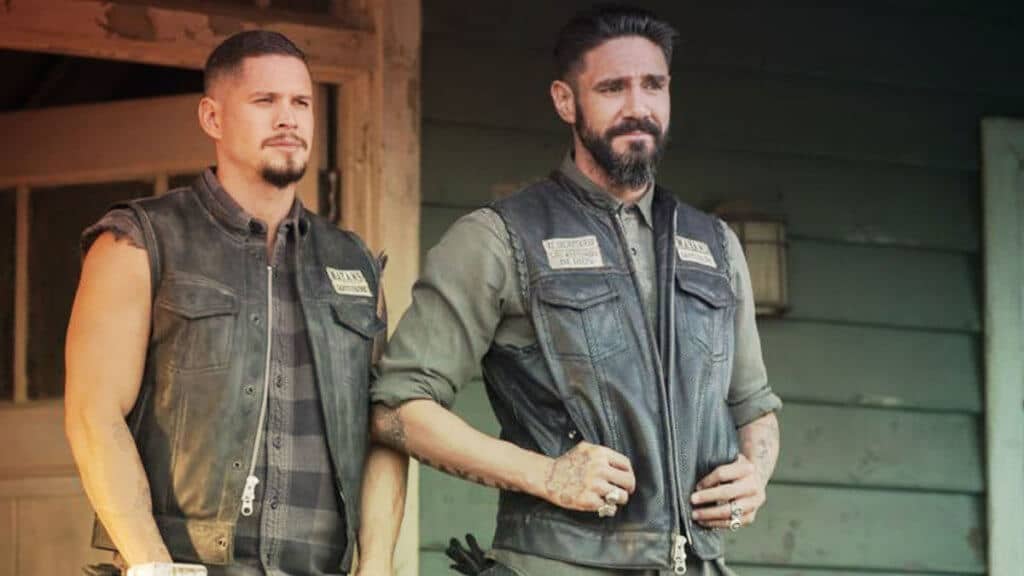 Two men in motorcycle vests stand on a porch