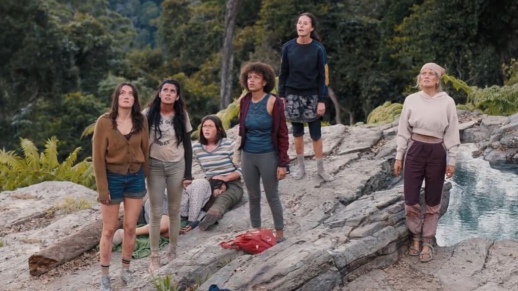 A group of tattered teen girls stand on a rock looking up
