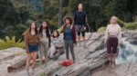 A group of tattered teen girls stand on a rock looking up
