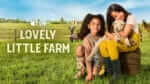 two young girls sit on a farm yard, snuggling a lamb