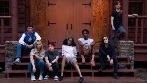 A group of queer teens on the steps of a cabin