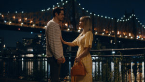 A couple standing by the river with a bridge lit up at night behind them