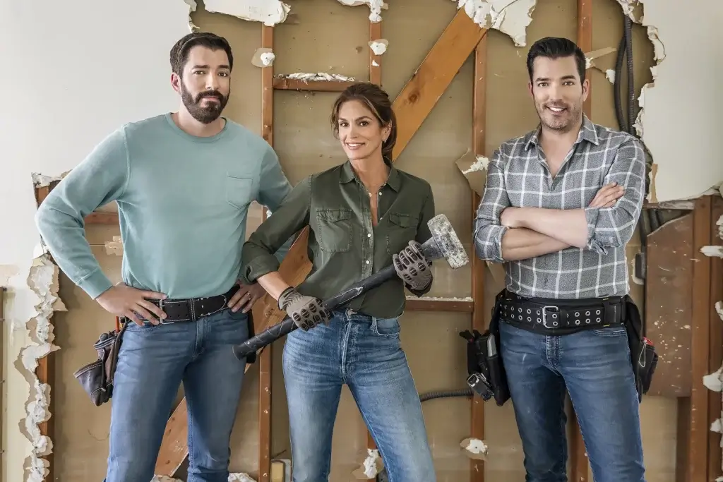 Two builders and a celebrity stand in a partially demolished home.