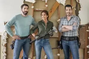 Two builders and a celebrity stand in a partially demolished home.