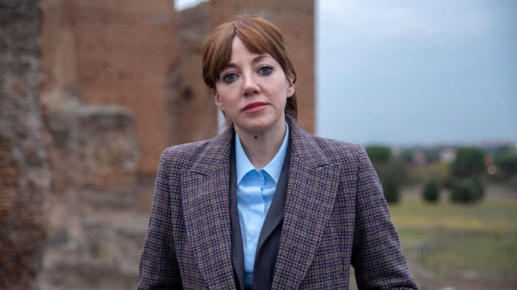 A woman in professional attire stands in front of historic ruins.