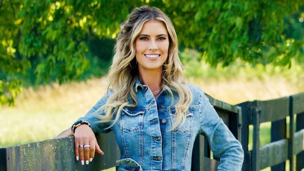 A blonde woman in denim stands by a farm fence