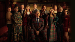 A group of reality star competitors in a dark manor room surrounding a host in plaid on a big leather chair