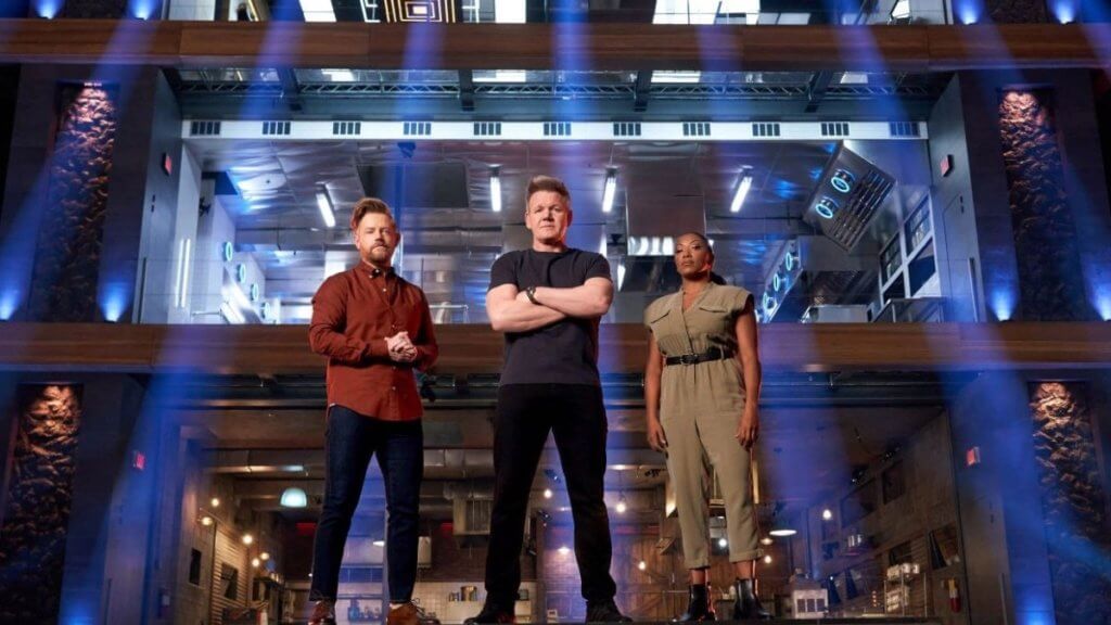 Three chefs stand under dramatic lighting in a competition kitchen arena