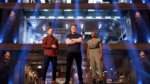 Three chefs stand under dramatic lighting in a competition kitchen arena