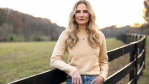 A blonde woman stands against a farm fence by rolling hills and pastures