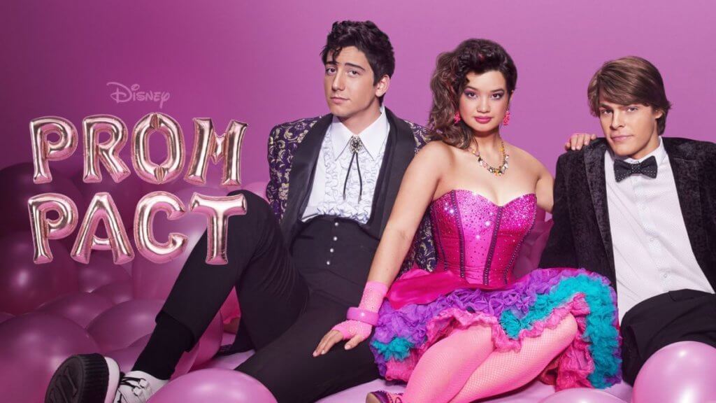 Three teens in 80s prom attire sit surrounded by pink balloons