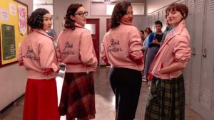 Four girls in 1950s clothes looking over shoulders at camera wearing pink jackets with Pink Ladies logo