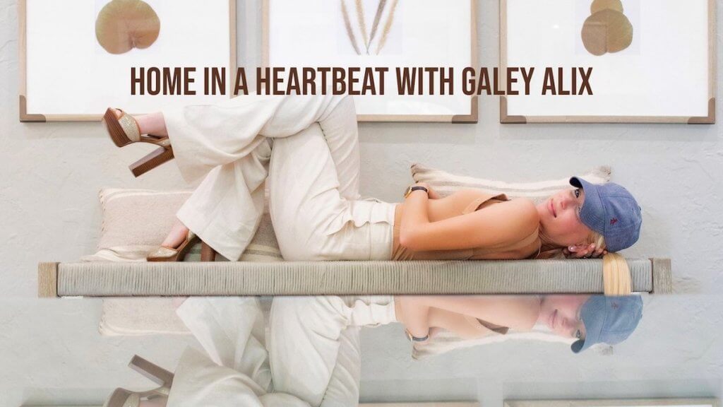 A stylish blonde woman in a baseball cap lays on a reflective table top turning to look at the camera