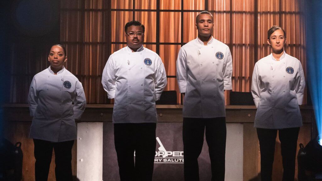 Four chefs in white jackets stand in military at ease with arms behind backs.