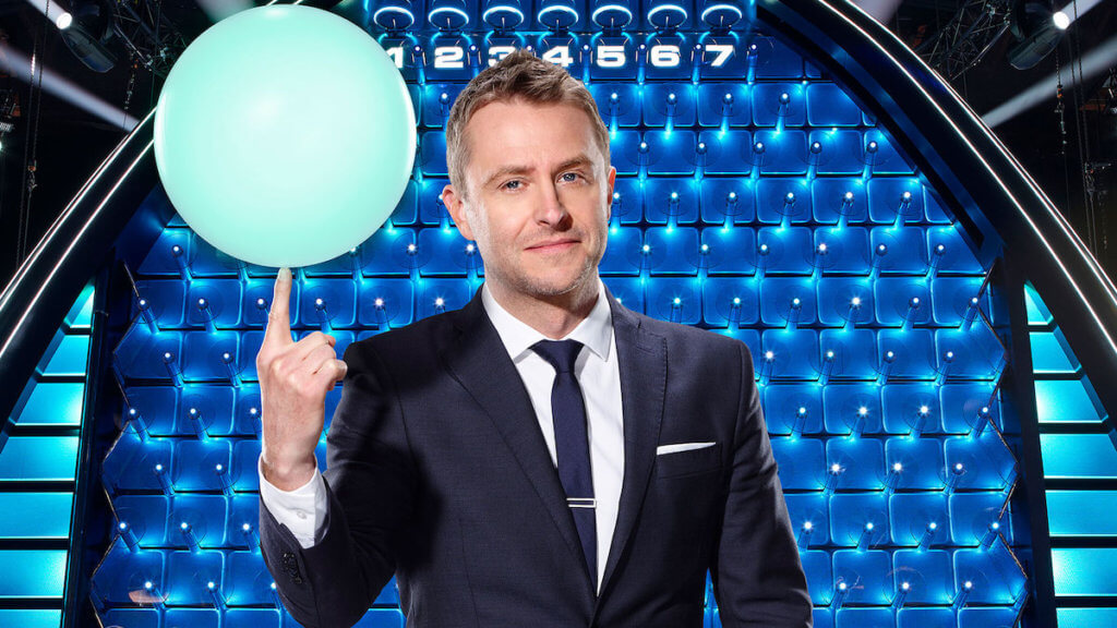 Host Chris Harwick balancing a blue glowing ball on one finger in front of a lit-up game board