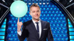Host Chris Harwick balancing a blue glowing ball on one finger in front of a lit-up game board