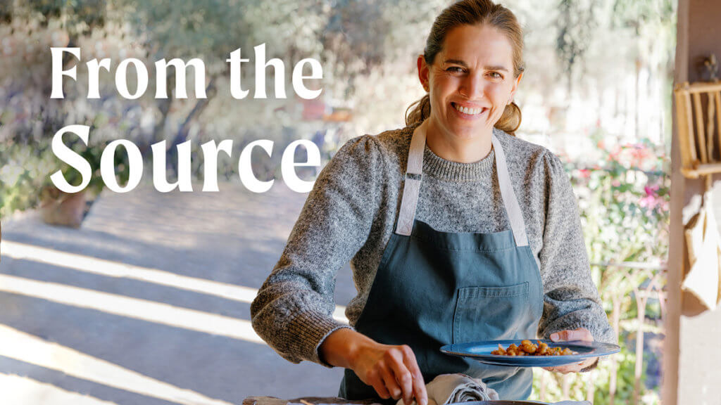 A woman chef in a denim apron sprinkling something in a pan with an open outdoor area behind her.