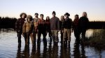 A group of 10 people in wilderness gear stand in a remote stream