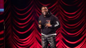 Comedian Kevin Hart in front of a red draped curtain on stage.