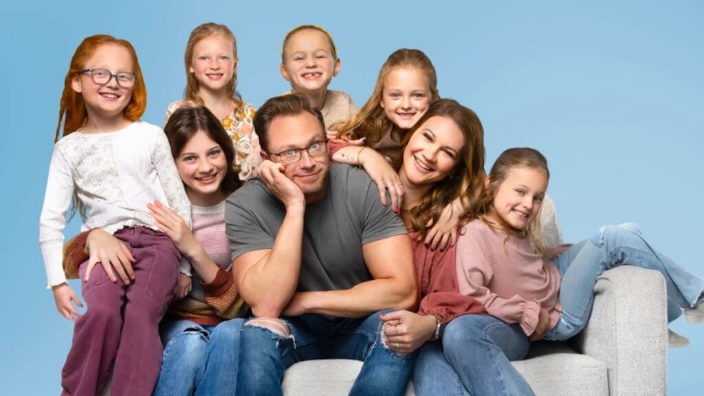 A couple and their older daughter sit in a chair with their five other daughters surrounding and leaning on them