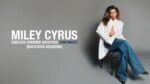 Singe Miley Cyrus sits on a chair in a blank grey background, wearing an oversized white shirt and jeans