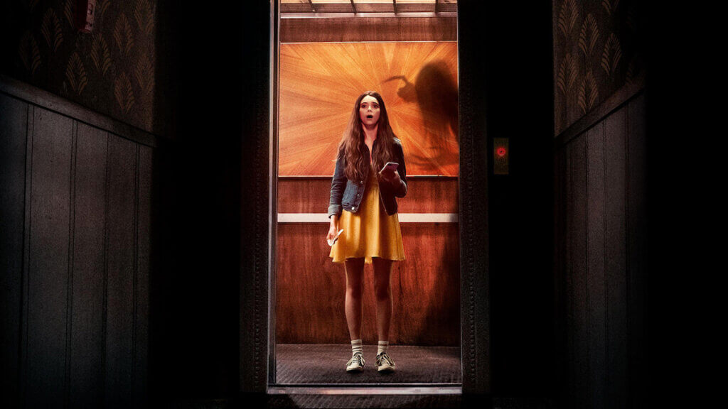 A young woman stands in an elevator facing a log dark hallway with an eerie shadow behind her.