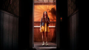 A young woman stands in an elevator facing a log dark hallway with an eerie shadow behind her.