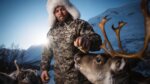 A man in winter gear and fur-lined hat feeding reindeer with snowy mountains behind him.