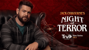 Jack Osbourne sits on a large leather chair looking at the camera in front of a deep red background with spider webs