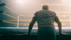 A muscular man leans on the edge of a wrestling ring with his back to the camera.
