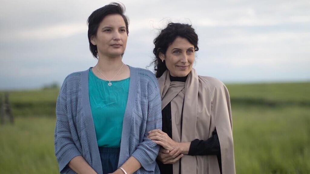 A young indigenous woman stands arm in arm with an older Jewish woman in a wide open prairie