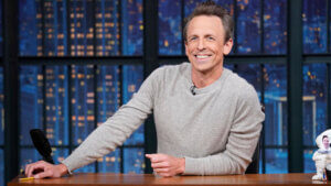 Late Night host Seth Meyers leans on his desk in a grey sweater